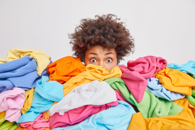 Shocked woman with curly Afro hair stares bugged eyes drowned in huge pile of colorful clothing cleans out closet selects clothes for donation or recycling white background. Housework concept